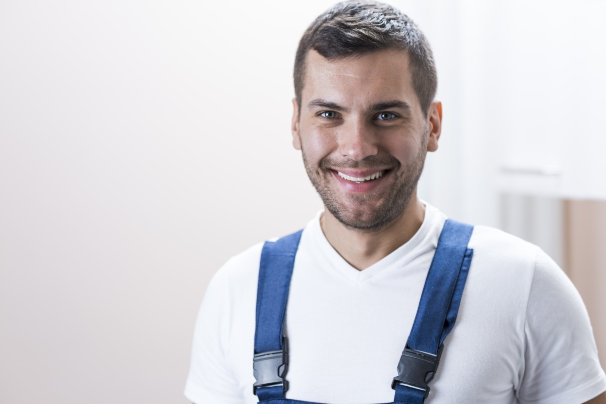 Junger Handwerker mit blauen Latzhosen und weißem T-Shirt, der in die Kamera lächelt.